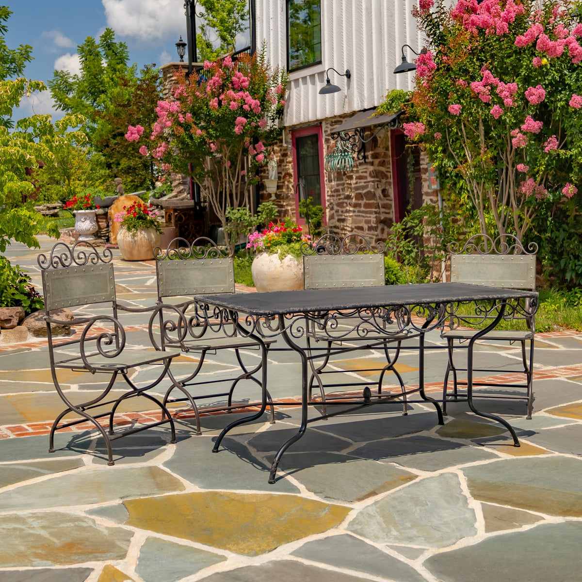Metal dining table and four matching chairs arranged on a stone patio among flowering shrubs.