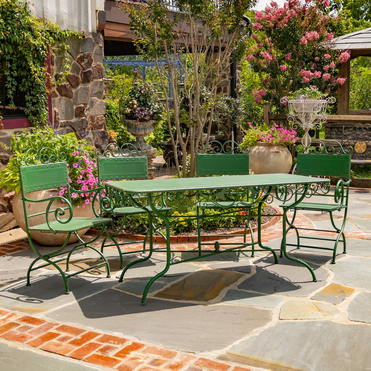 Green metal dining table and four ornate chairs arranged on a stone patio amid flowering garden plants.