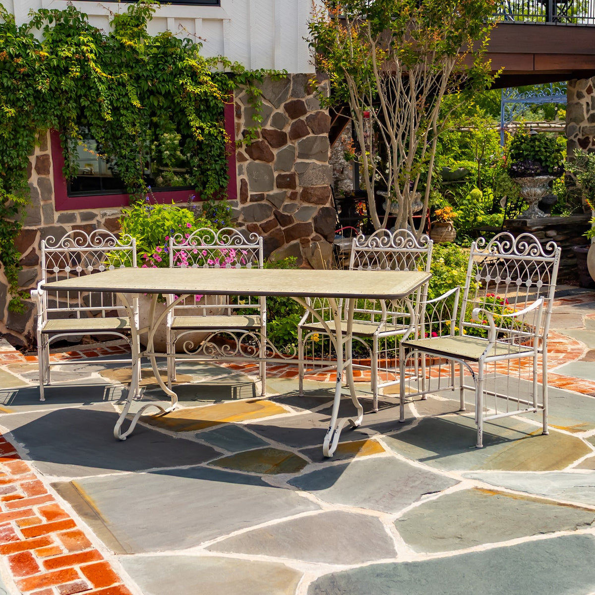 White metal dining table and ornate matching chairs arranged on a stone patio among garden greenery.