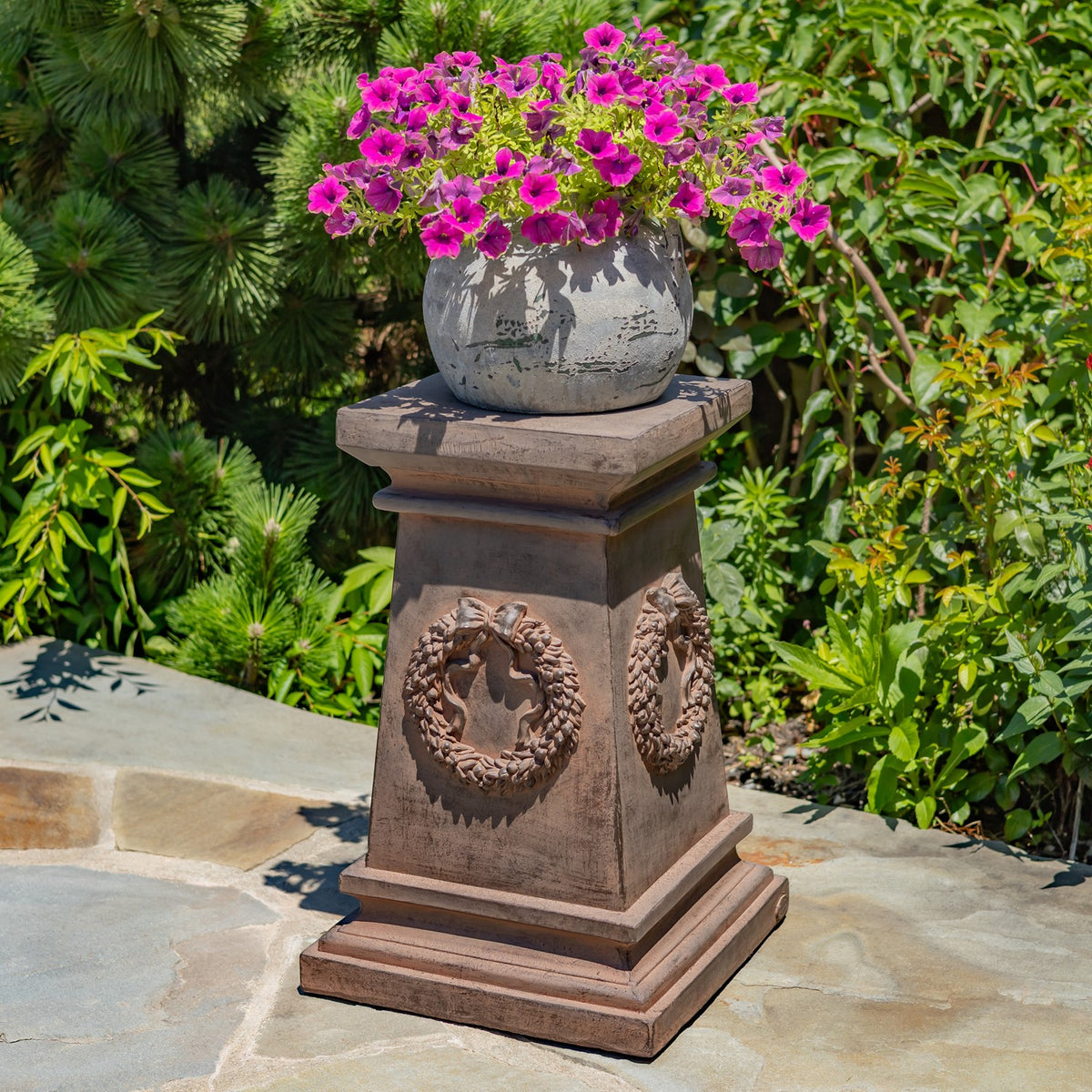Brown decorative column with wreath reliefs beneath a flowerpot of pink flowers, surrounded by greenery.
