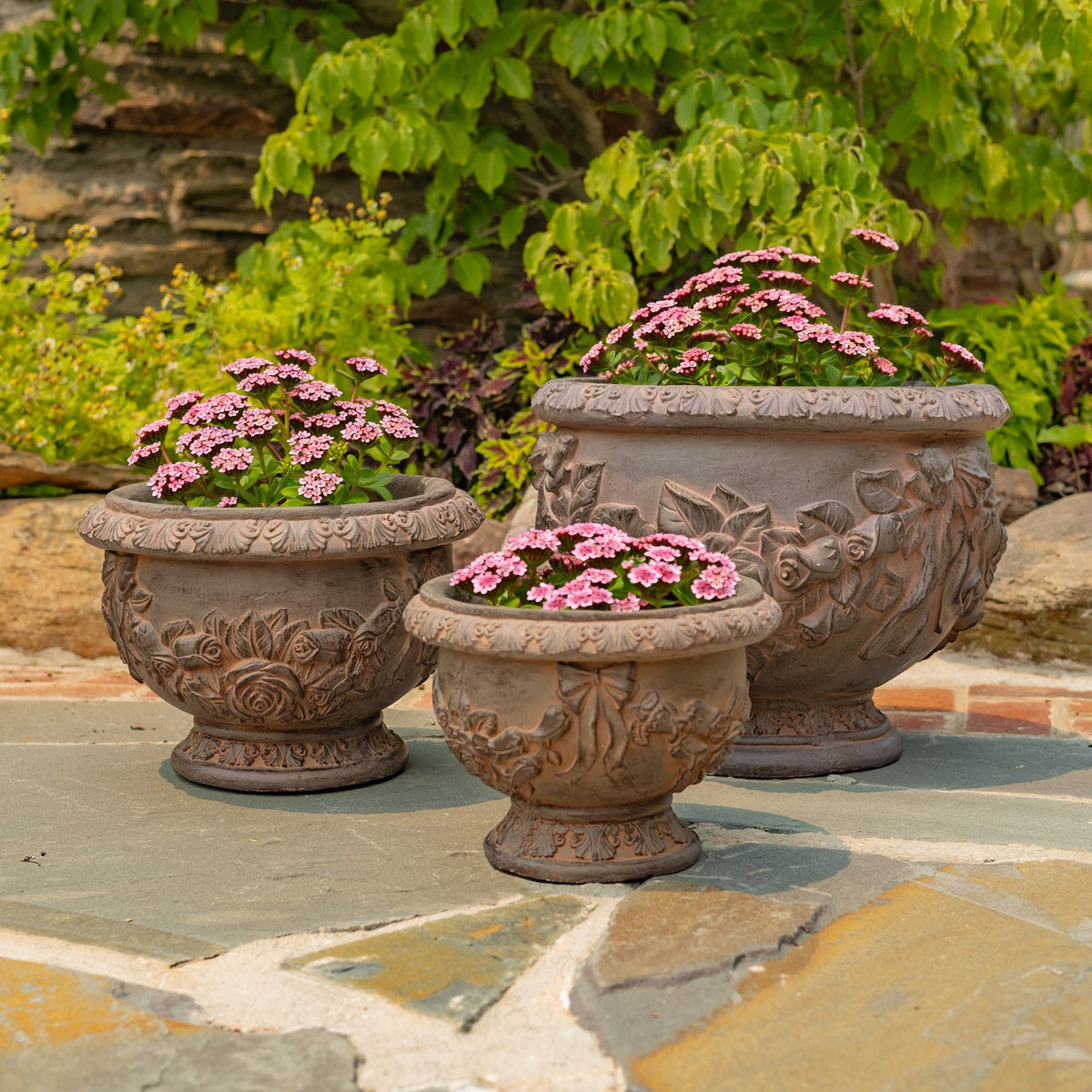Three ornate rust-toned round planters with pink flowers arranged on a stone patio.