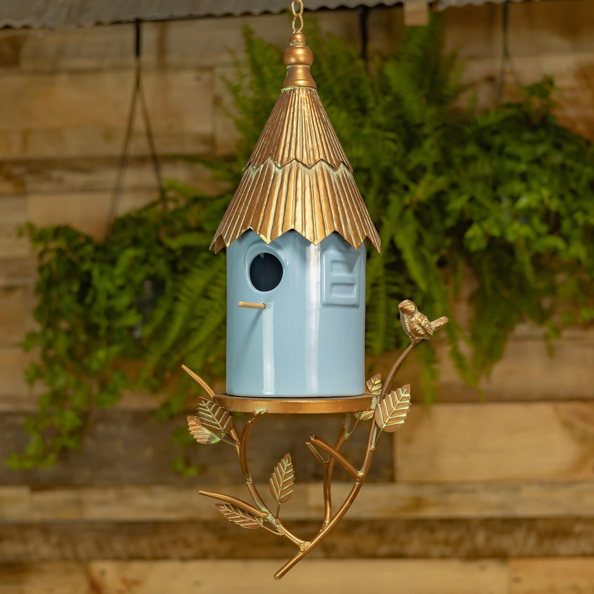 Hanging blue birdhouse with a gold-toned roof and branch-shaped frame, displayed against a leafy garden backdrop.
