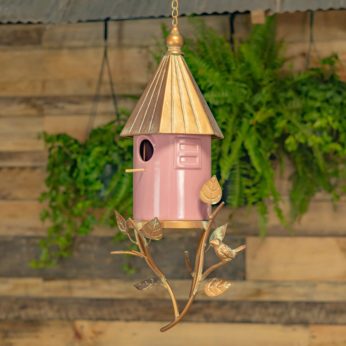 Pink birdhouse with a gold-toned roof and branch-shaped hanger, suspended against a wood and greenery backdrop.