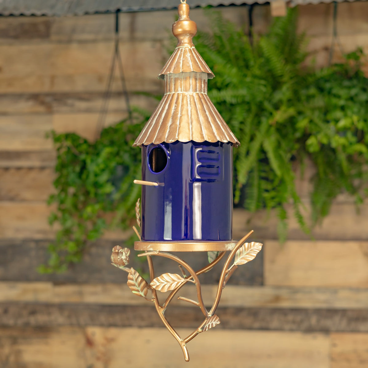 Hanging birdhouse with a cobalt blue body, copper-colored roof, and decorative branch stand against a garden backdrop.