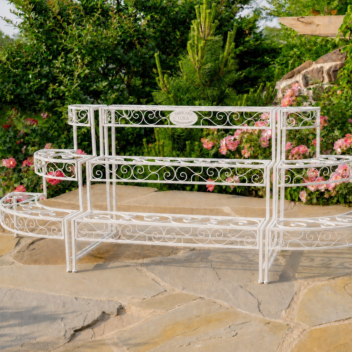 Antique-white three-tier metal plant stand with ornate scrollwork on a stone patio among flowering shrubs.
