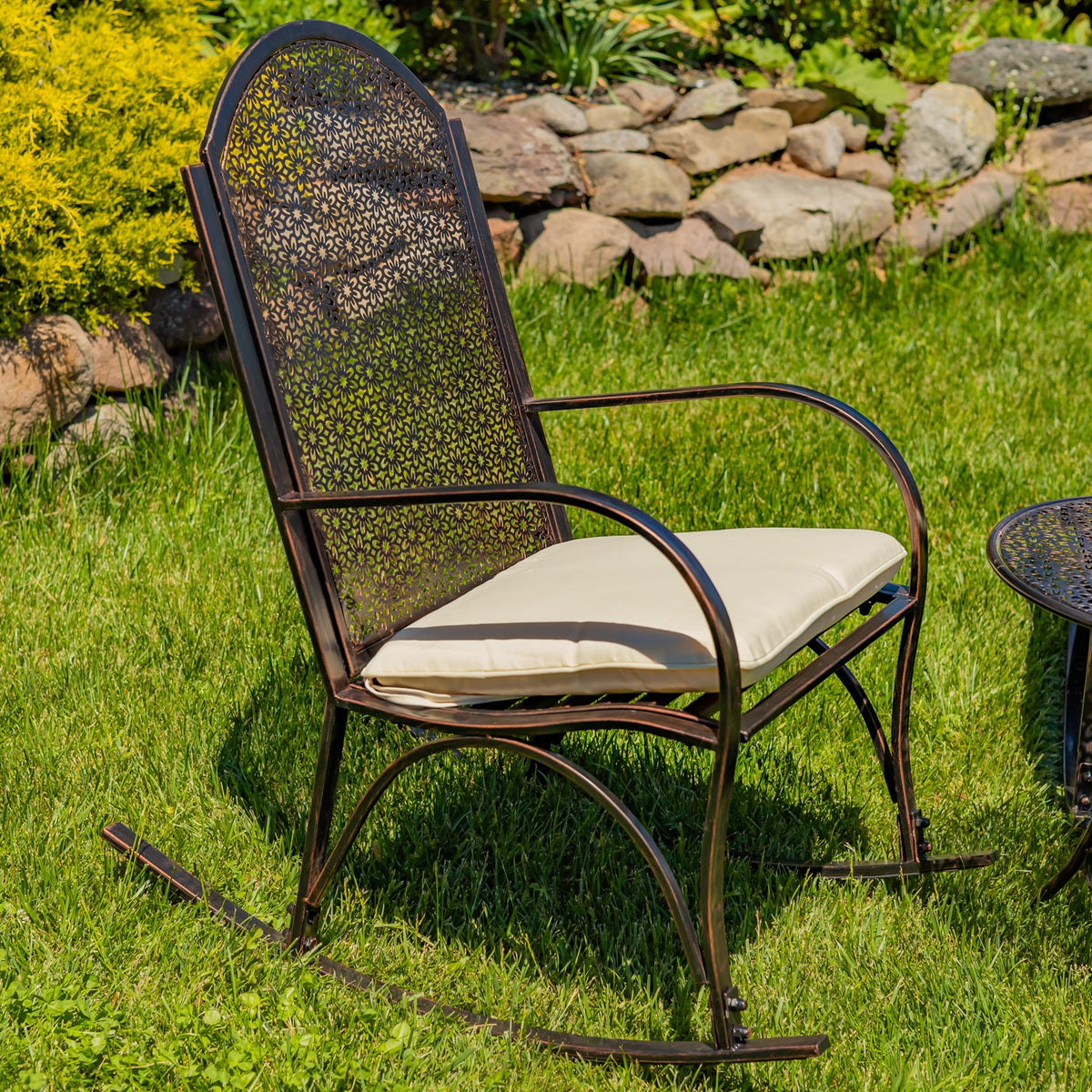 Ornate dark metal rocking armchair with a light cushion on a lawn beside a stone garden border.