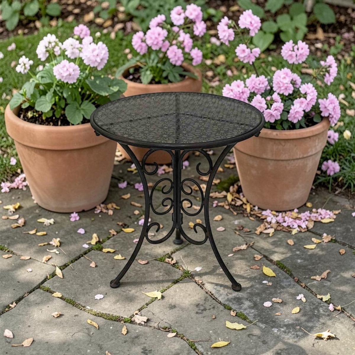 Round dark metal side table with a patterned top and curled legs on a stone patio among flower pots.