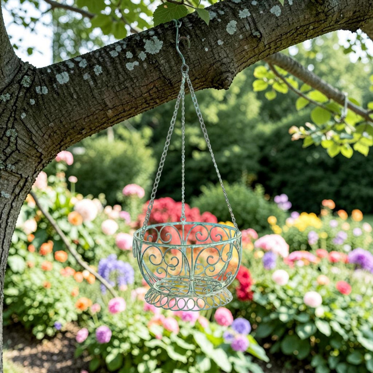 Antique green scrollwork hanging basket suspended from a tree branch above a colorful flower garden.