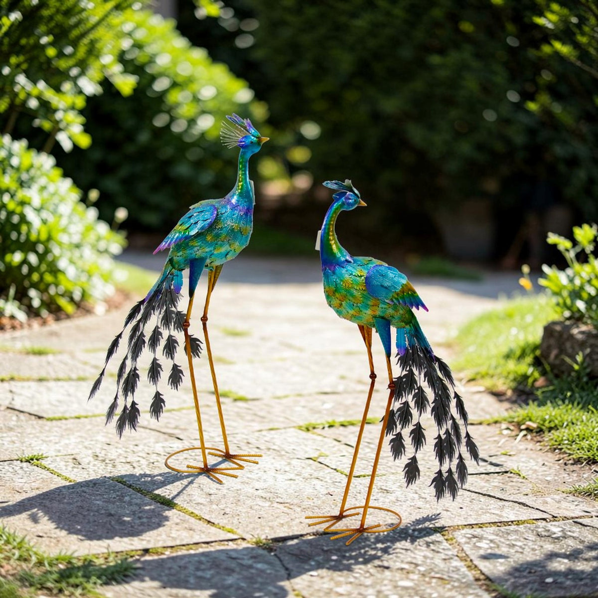 Pair of blue and green metallic peacock sculptures on a sunlit garden path