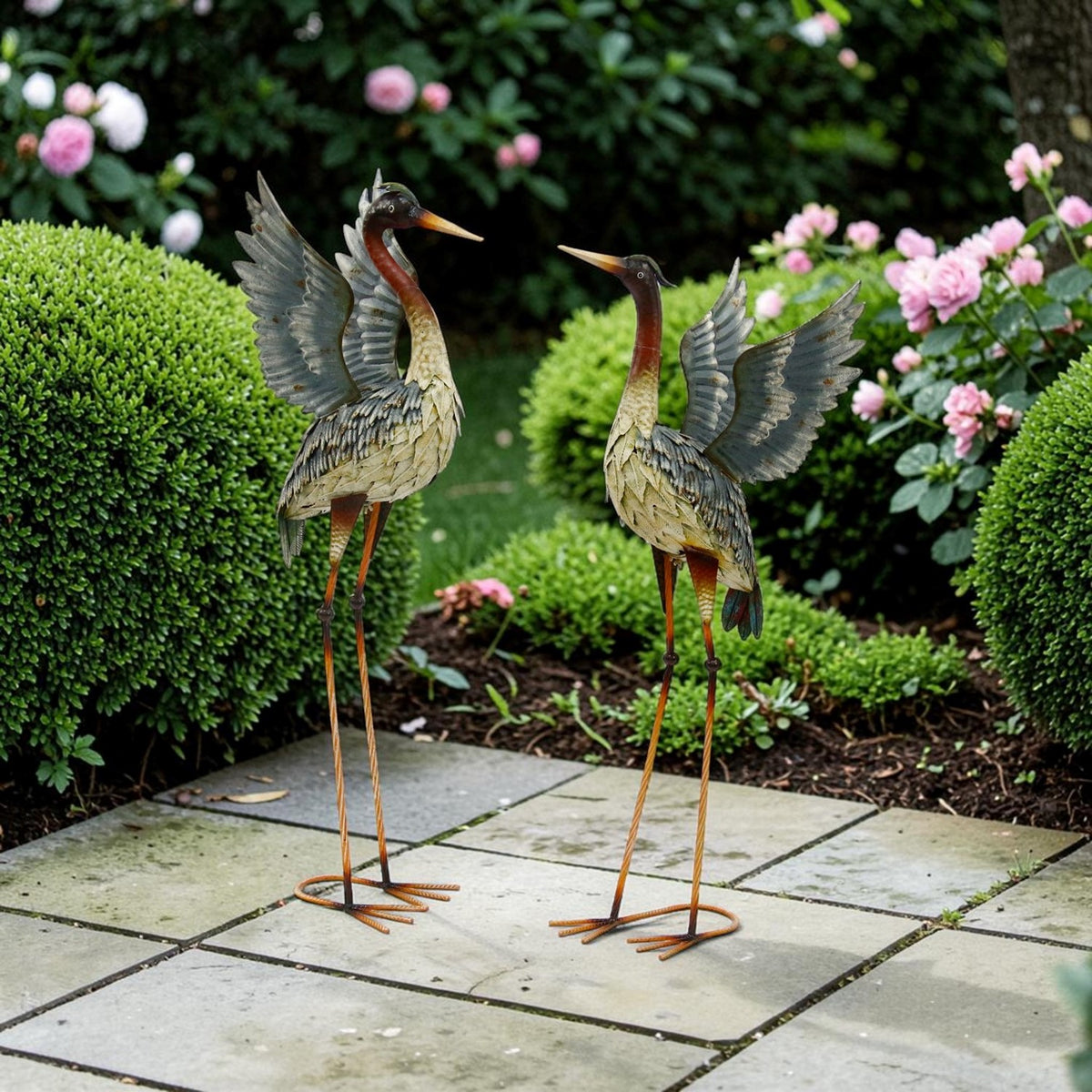 Two metal heron sculptures with raised wings stand on a stone patio among flowering garden plants.