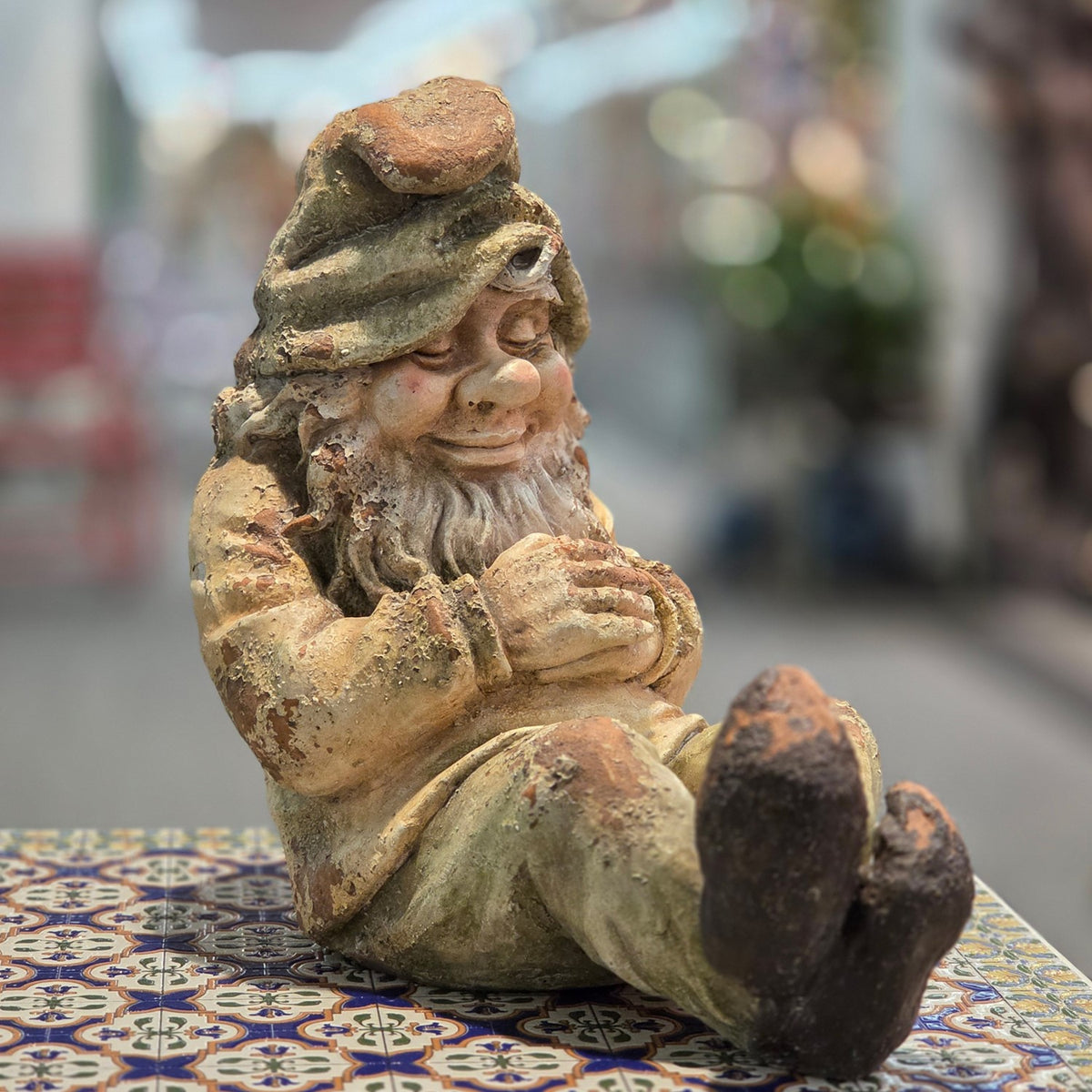 Distressed sleeping garden gnome seated on a patterned table, angled front view
