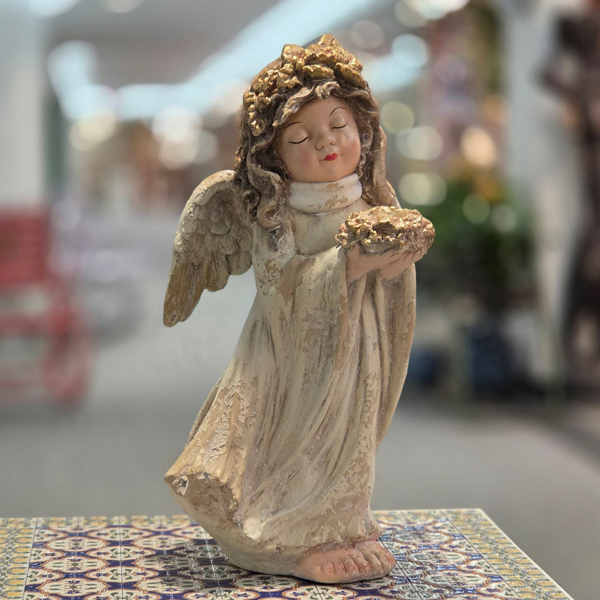 Distressed pastel angel figurine holding flowers, shown from the front on a patterned table