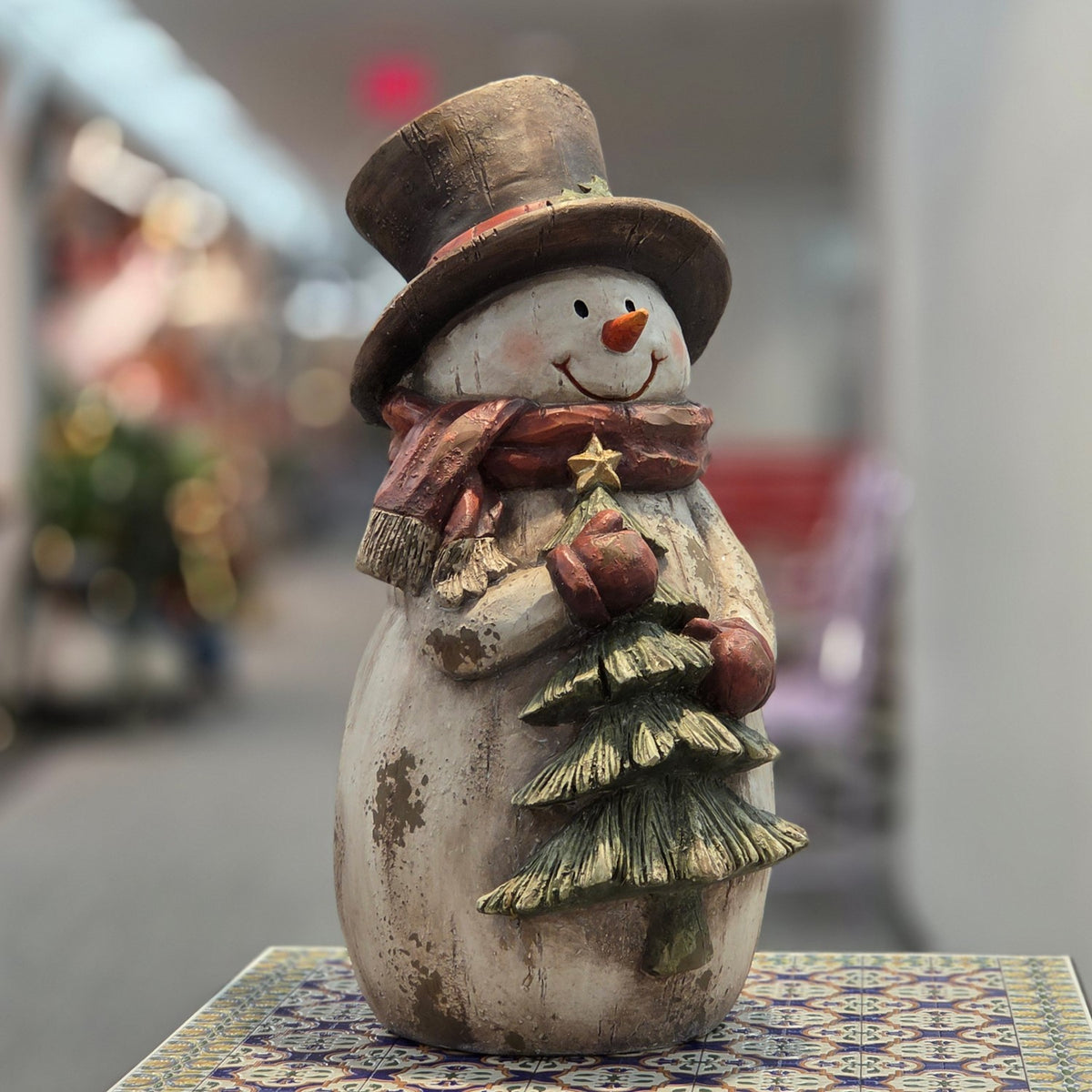Distressed snowman figurine holding a decorated tree, front view on a patterned table