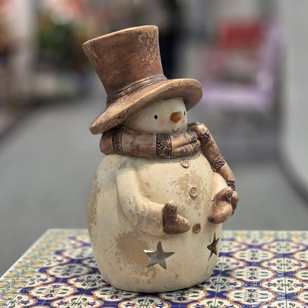 A cream-colored snowman candleholder with a brown top hat, scarf, and star cutouts stands on a patterned table.