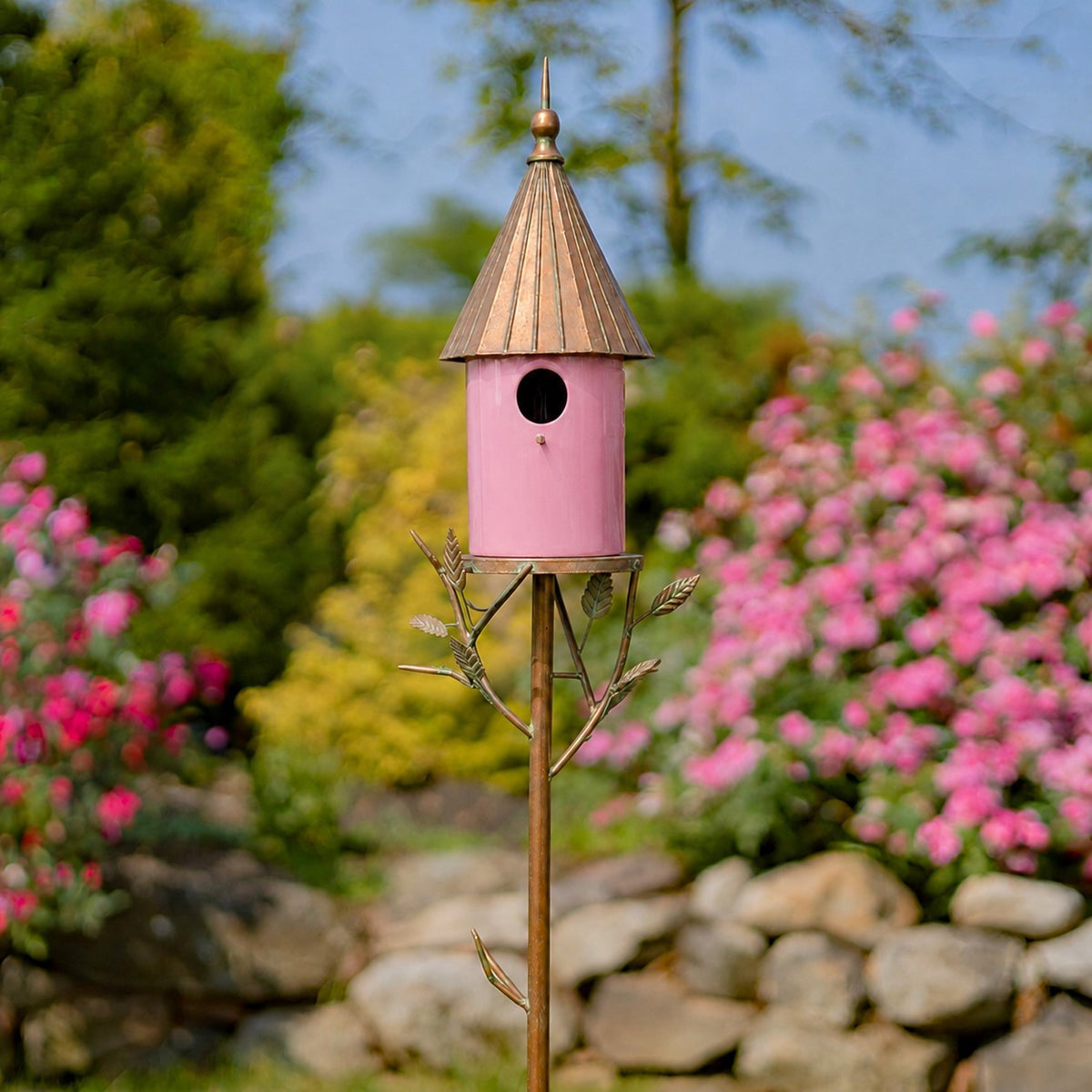 Pink birdhouse stake with a copper-toned roof stands among flowering shrubs and garden stones.