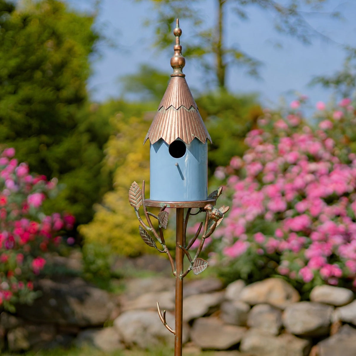 Blue birdhouse stake with a copper-toned roof and leafy metal branches among flowering garden plants.