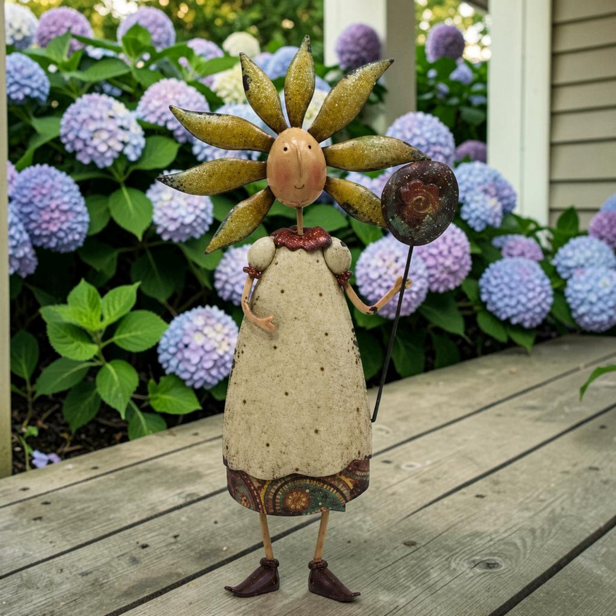 Metal flower-headed lady figurine standing on a wooden deck in front of blooming hydrangeas.
