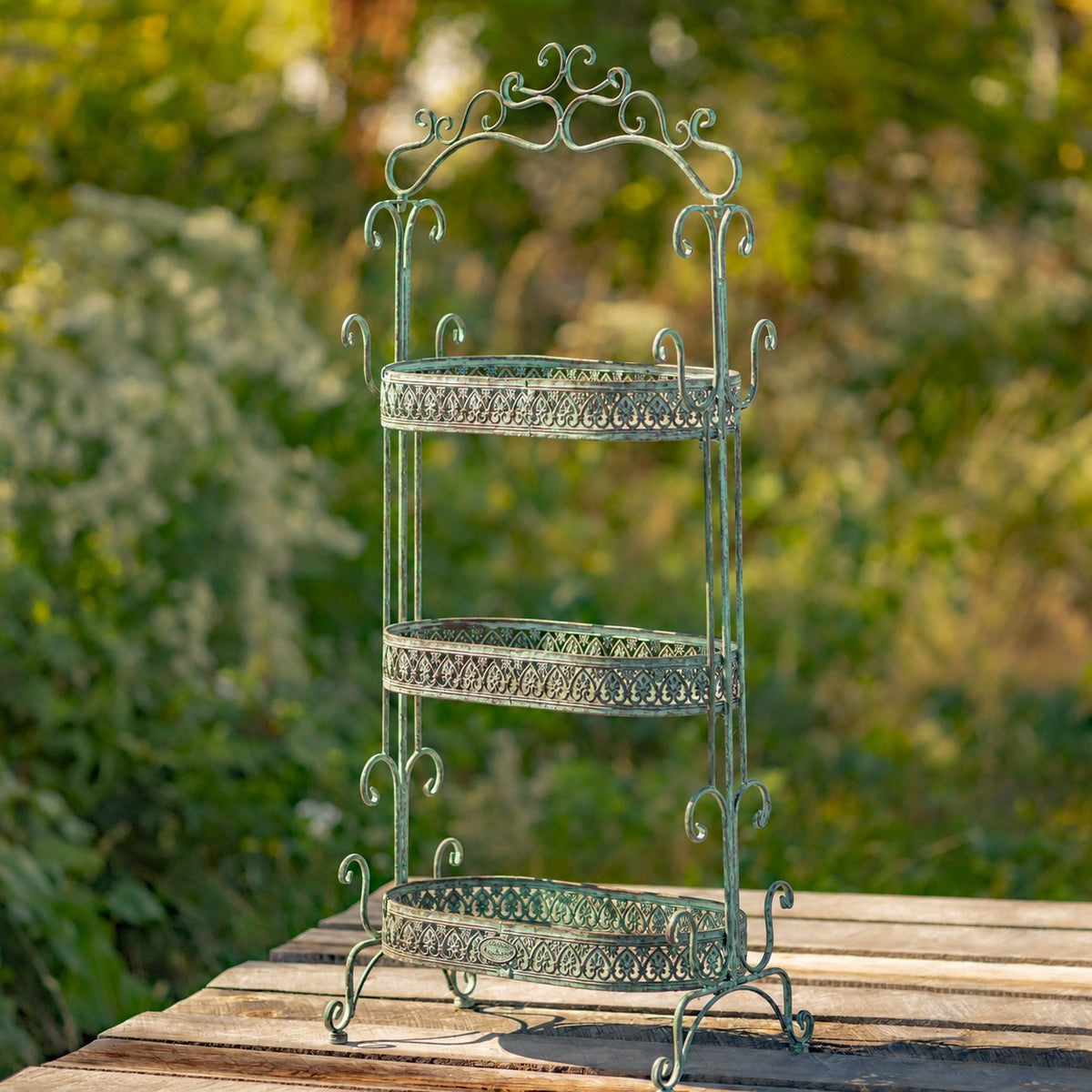 Ornate green iron plant stand with three empty tiers on a wooden outdoor deck.