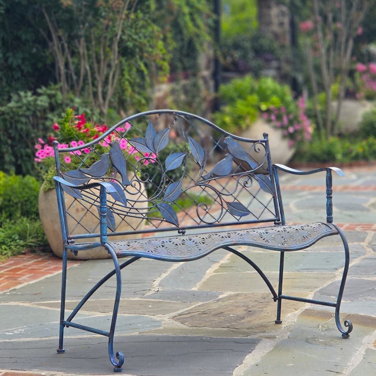 Blue metal garden bench with a leaf-patterned backrest on a stone patio among flowering plants.