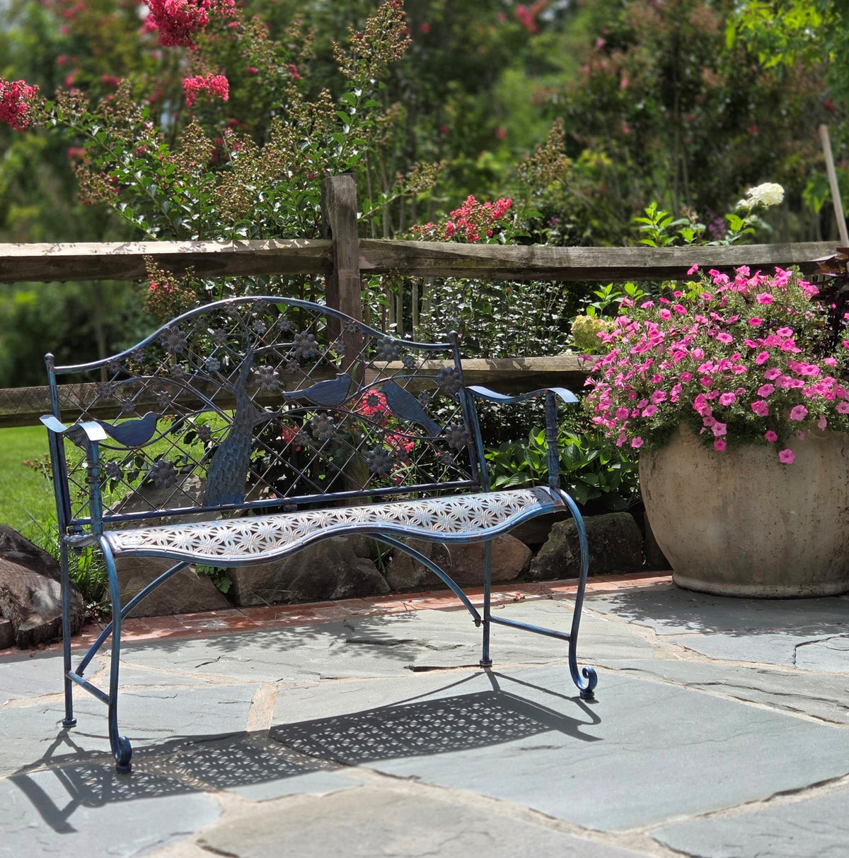Dark metal garden bench with a flowering tree and birds in its backrest beside a flower planter.