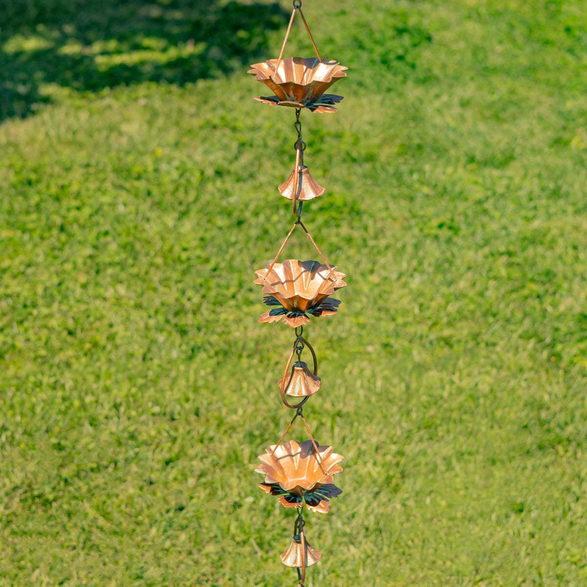 A copper-toned rain chain with lotus-shaped cups and small bells hangs against a green lawn.