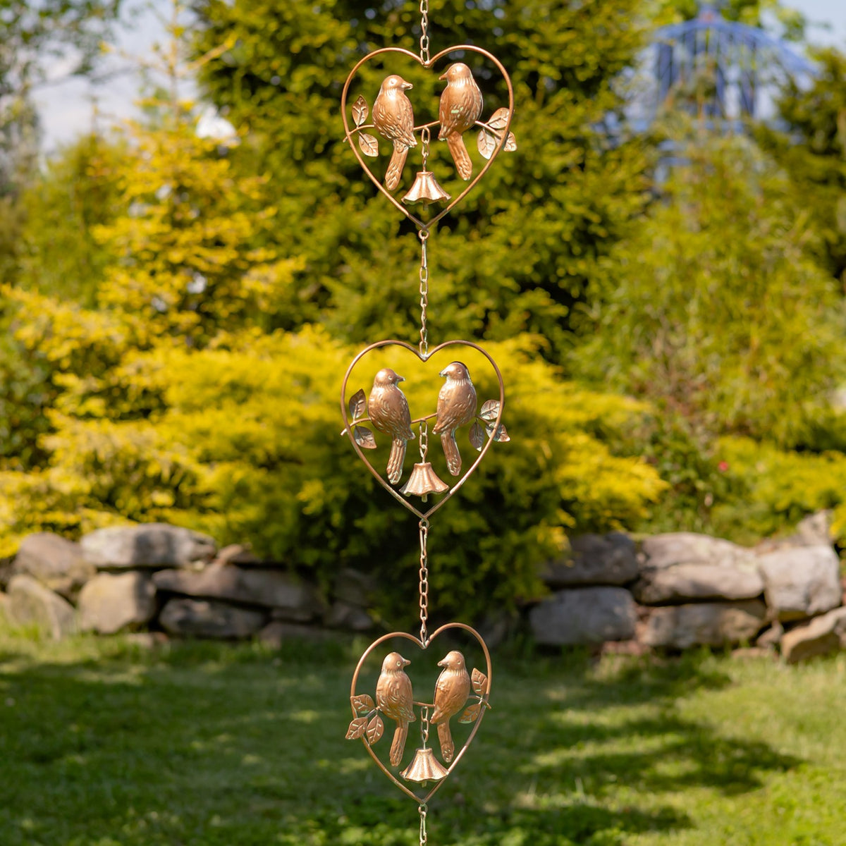 A vertical rain chain with three heart-shaped frames, paired birds and bells hangs against a garden backdrop.