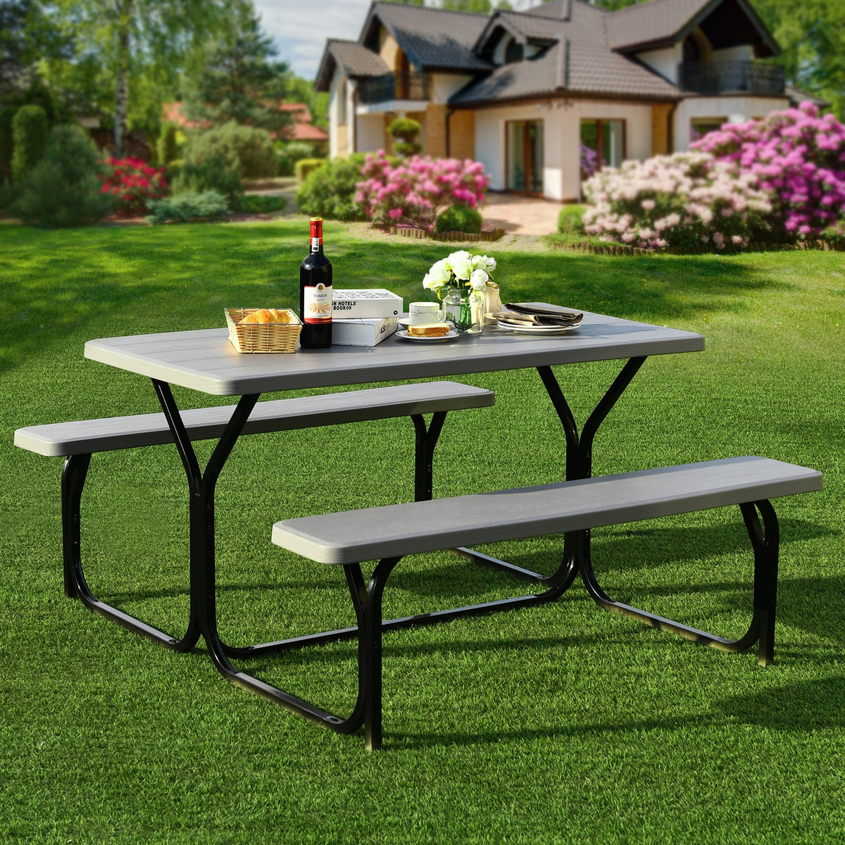 Gray picnic table and benches arranged on a lawn, set with dishes and a flower vase.