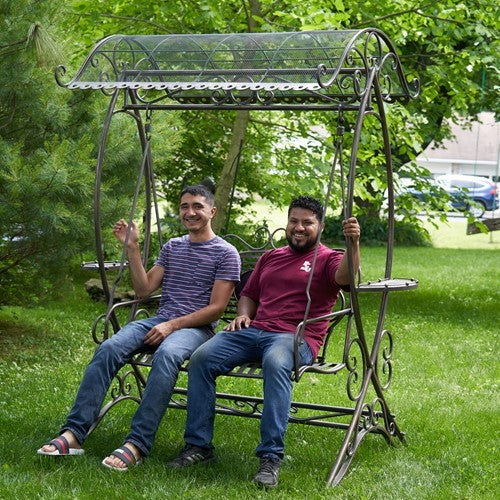 Two men sit on the ornate garden swing beneath its curved canopy on a grassy lawn.
