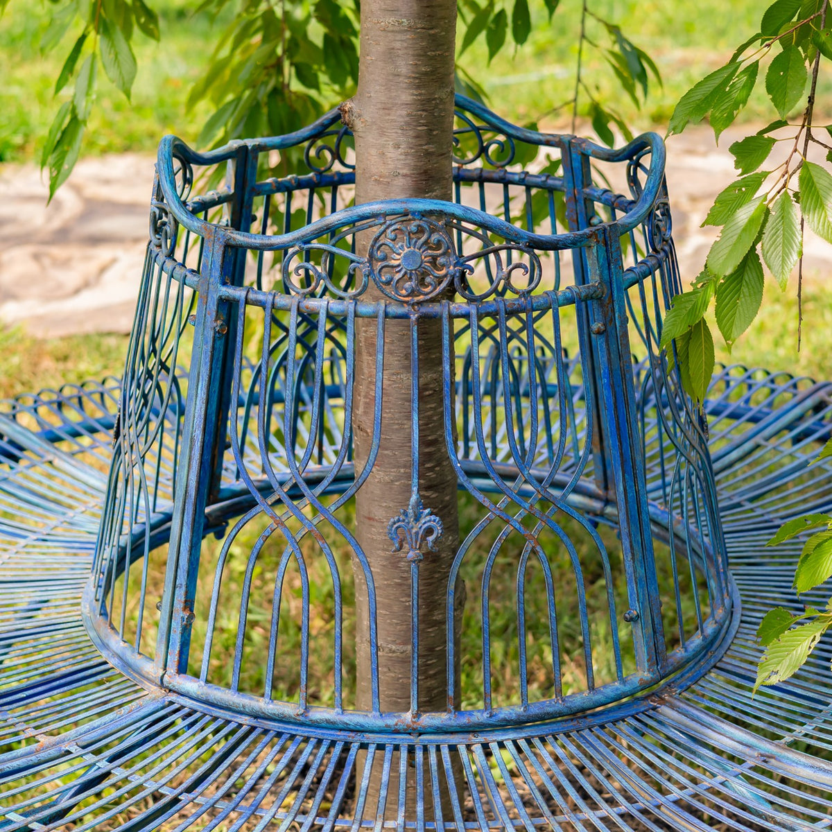 Blue metal tree bench encircling a tree trunk, with ornate scrollwork and a circular slatted seat.