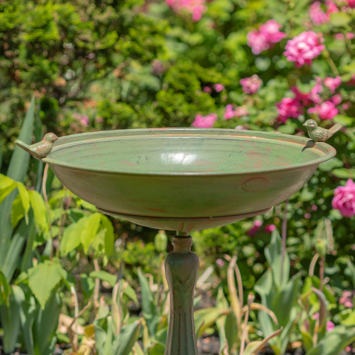 Green pedestal birdbath with small bird figures on the rim, set among flowers and foliage.