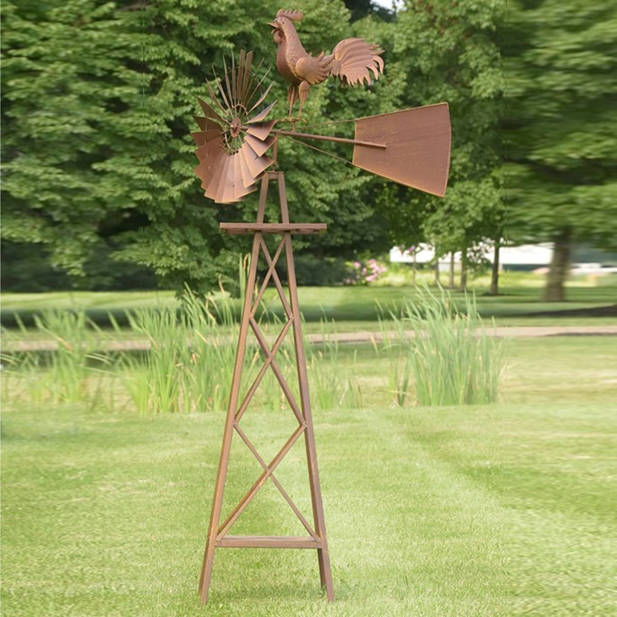 Tall cross-braced windmill stand with rooster, fan blades and tail vane against a garden backdrop.