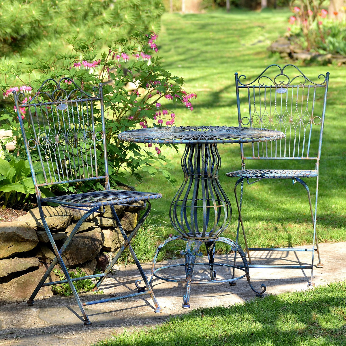 A round metal table and two folding chairs are arranged beside a flower bed on the lawn.