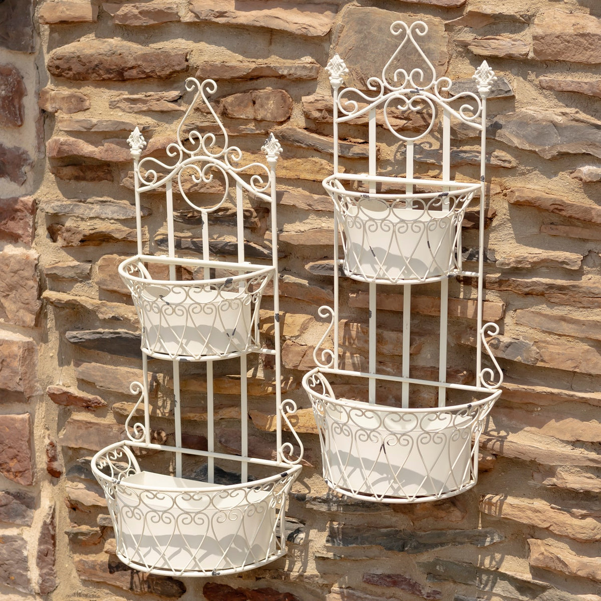 Two white wrought-style wall planters with empty removable baskets hang against a stone wall.