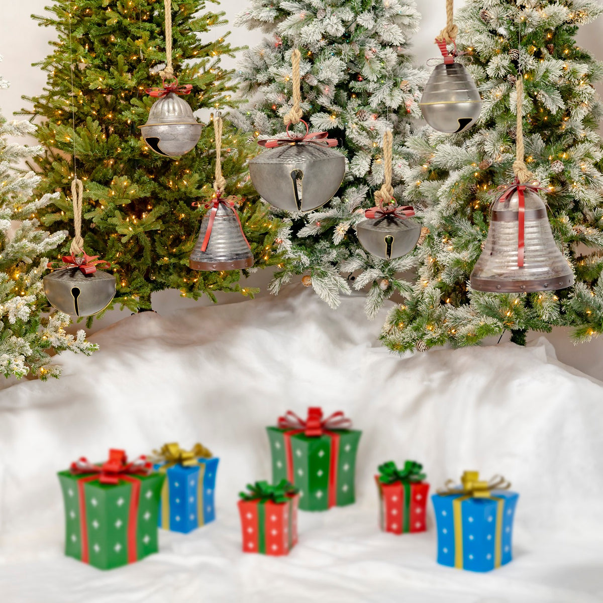Assorted metal bells with red ribbons hang on evergreen trees above colorful wrapped gifts.