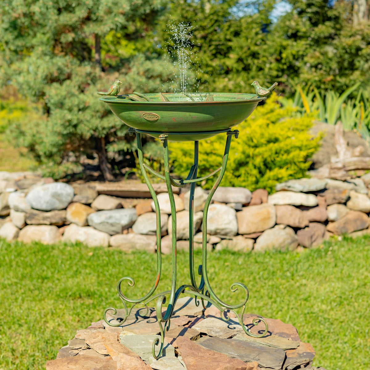 Full view of the birdbath on its curled metal stand, with water spraying from the bowl in a garden.