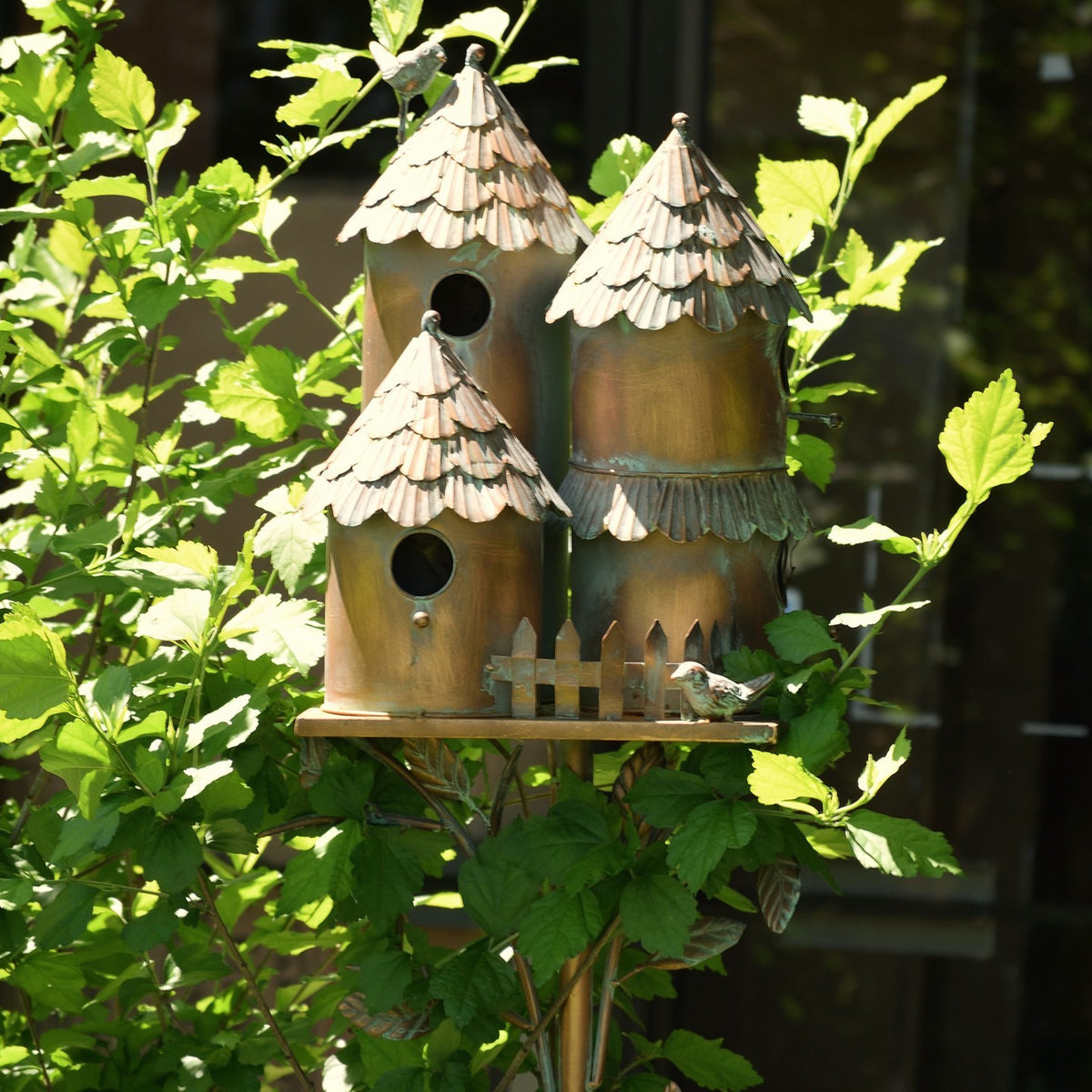 Three metal birdhouses and a small picket-fence detail are nestled among sunlit green leaves.