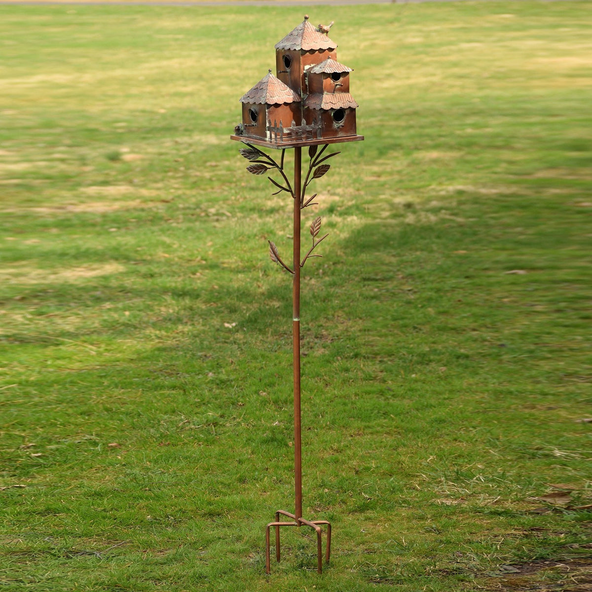 A tall metal birdhouse stake stands in a grassy yard, with a cluster of birdhouses above a leafy stem and ground prongs.