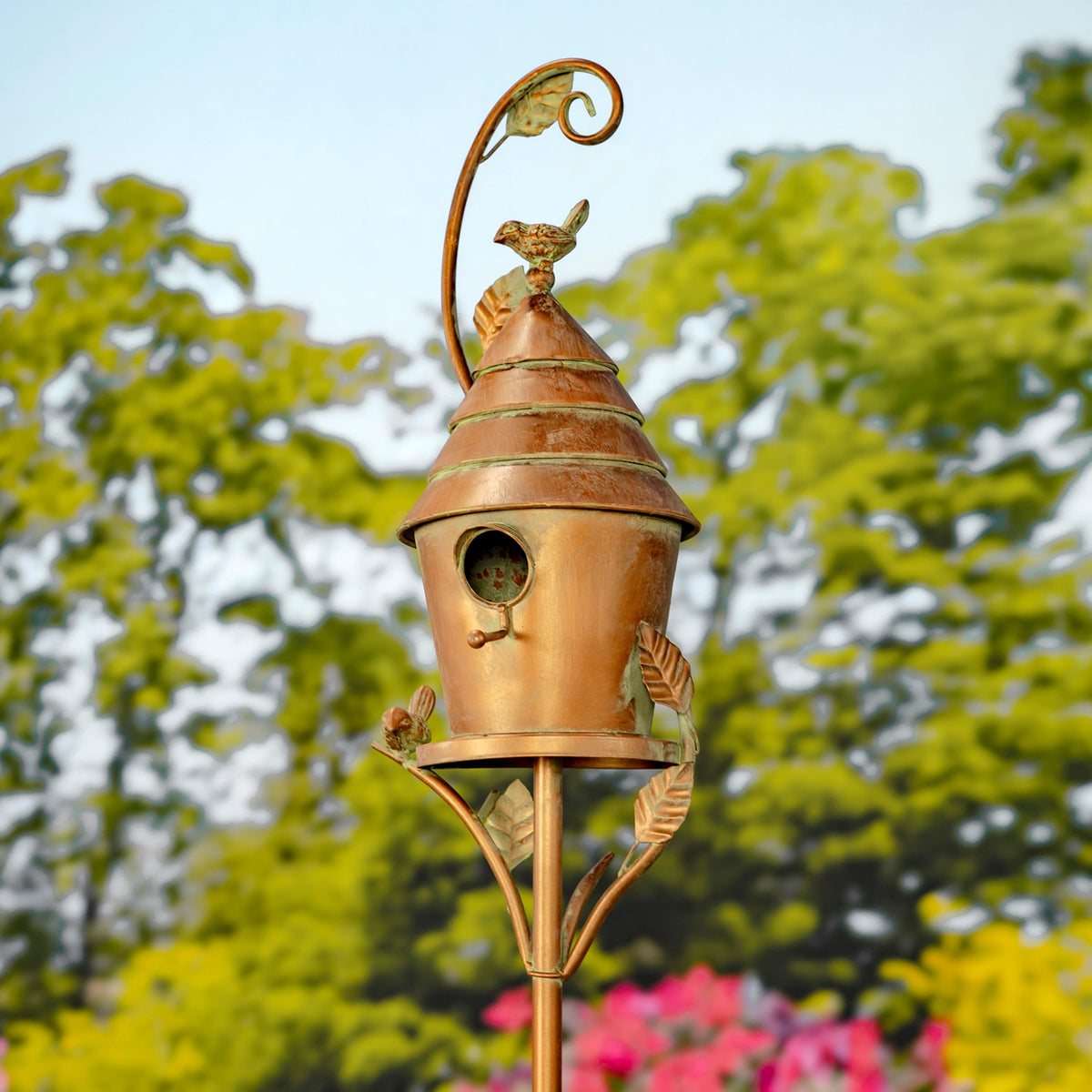 Full view of a copper birdhouse stake with curled metalwork, leaf details and a bird ornament against trees.