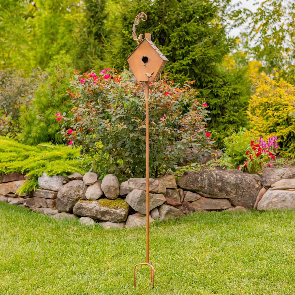 Full-height copper birdhouse stake planted in a lawn beside a stone border