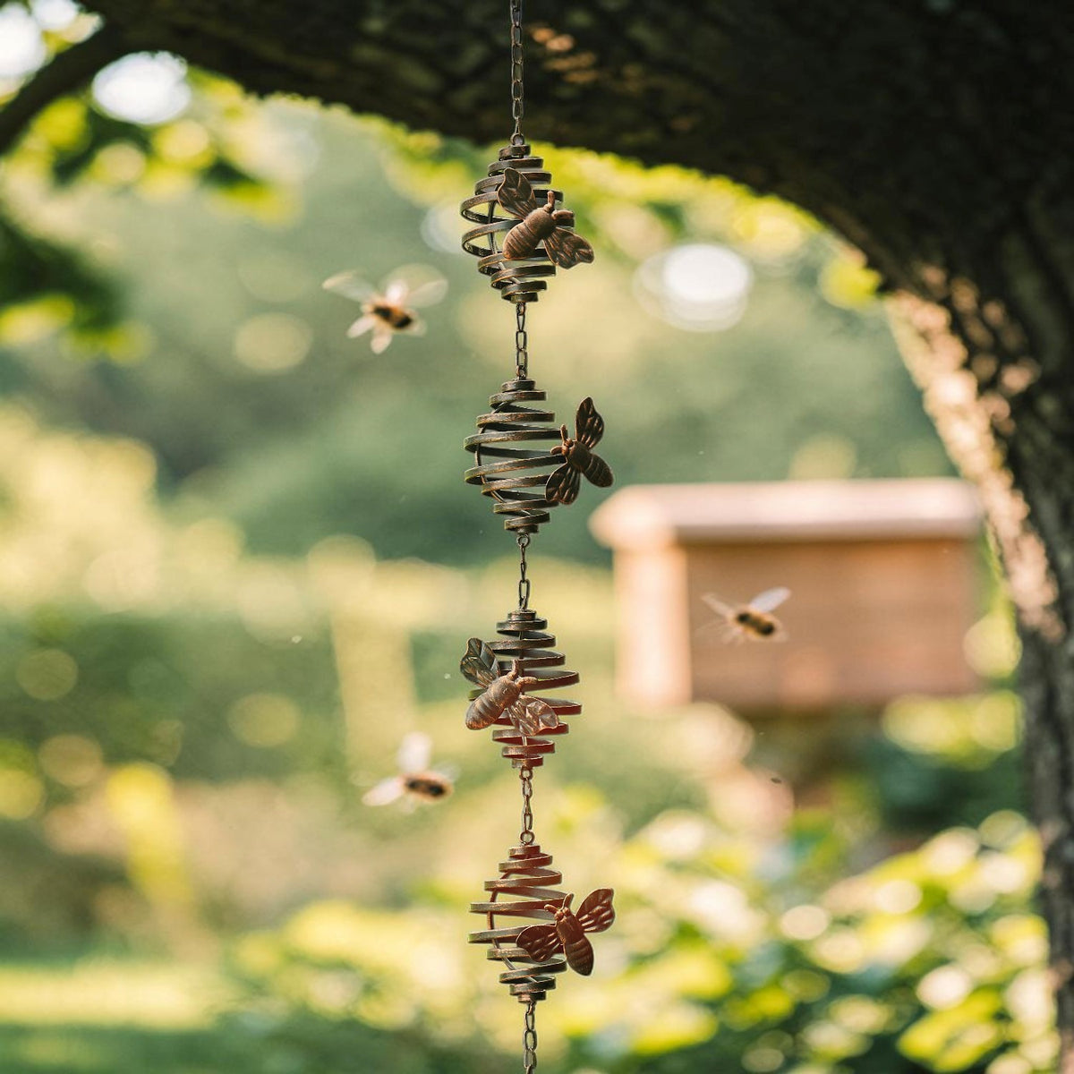 The bee-decorated rain chain hangs vertically from a tree branch against a softly blurred garden and birdhouse.