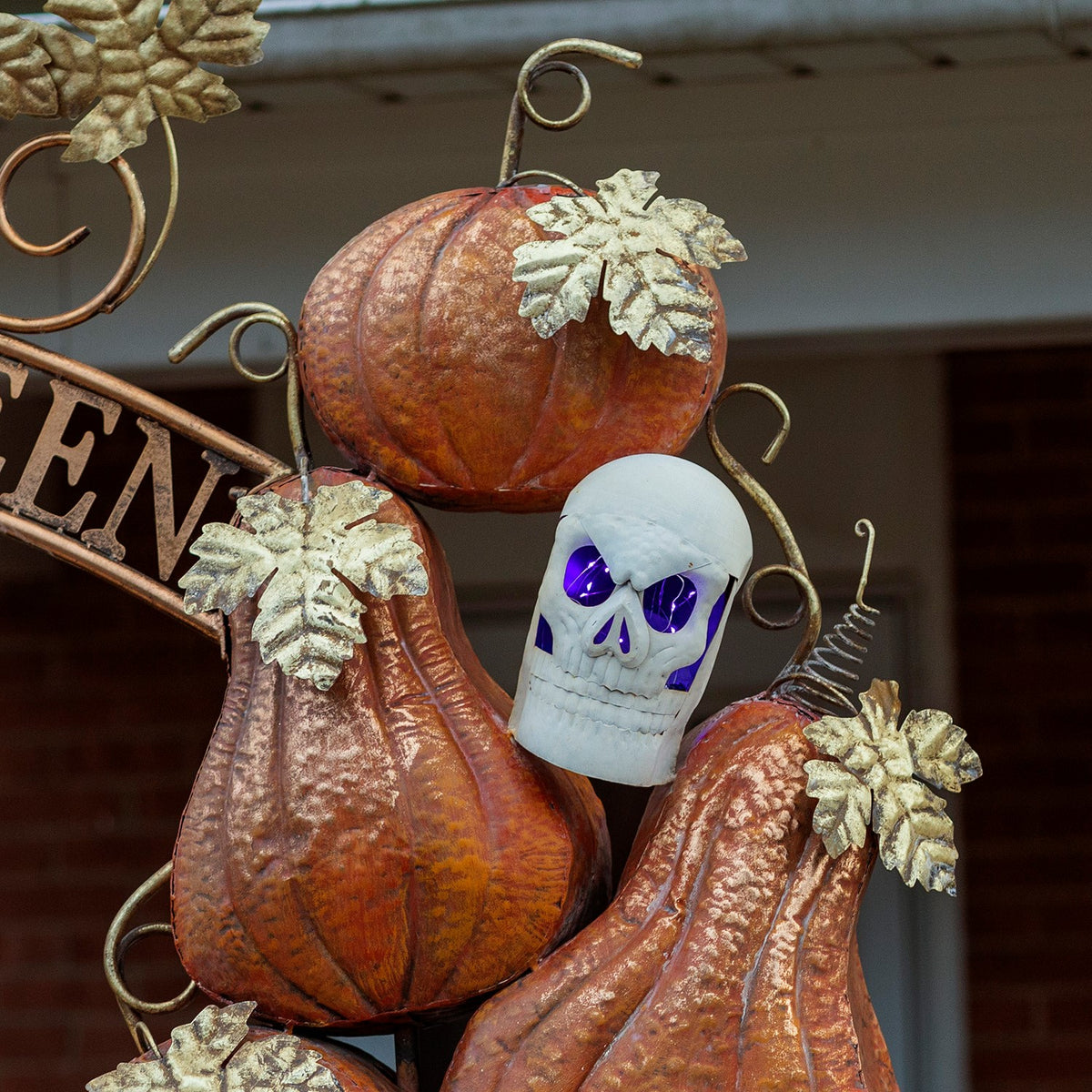 Close-up of textured orange pumpkins, pale leaves, curled metal stems, and a skull with purple-lit eyes.