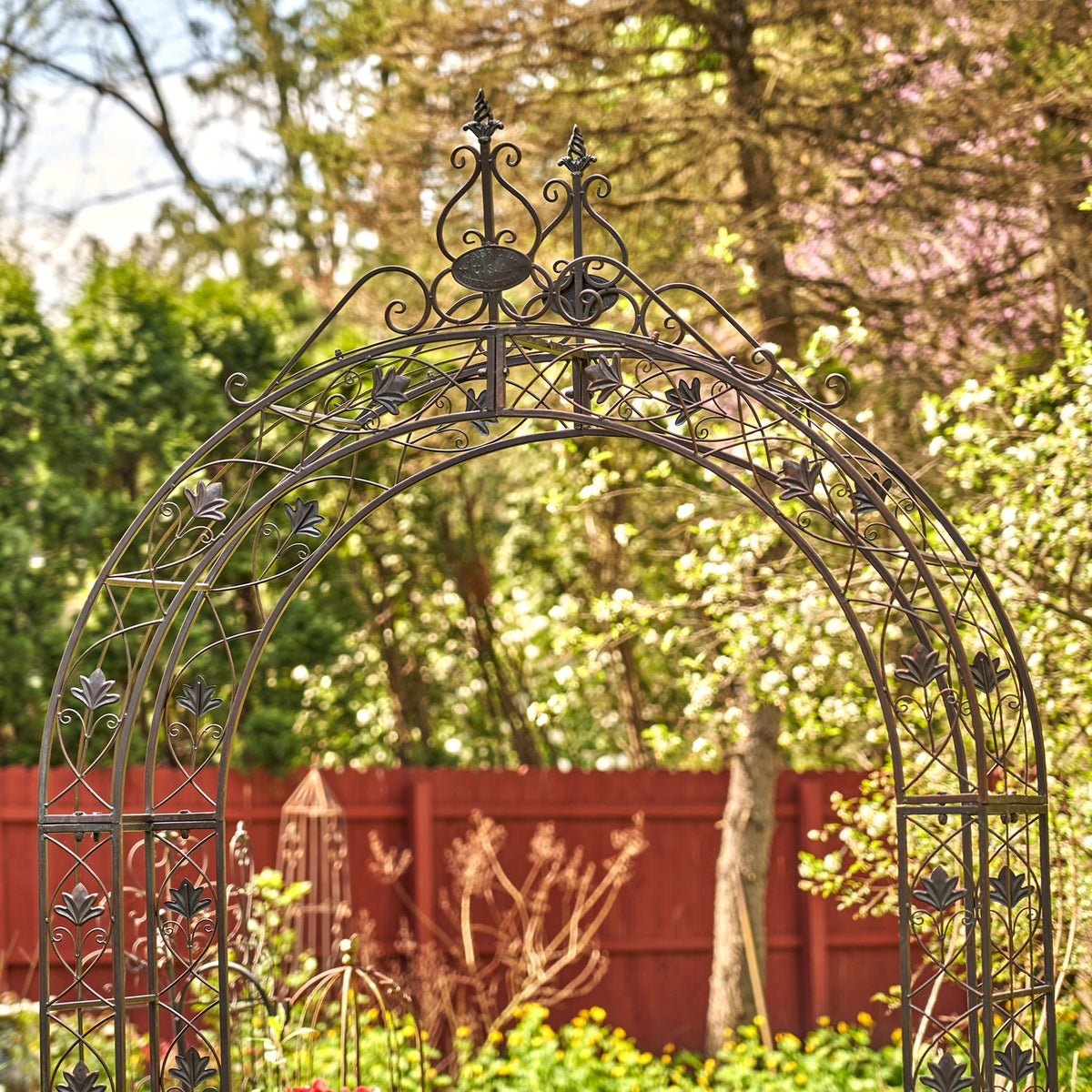 Dark metal arch with decorative scrollwork and leaf motifs against a sunlit garden.