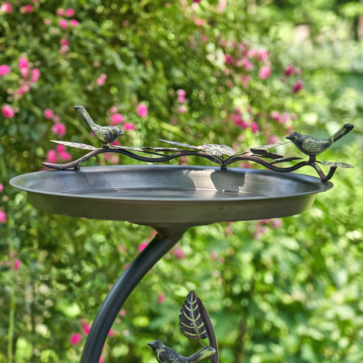 Shallow metal birdbath with a branch-and-bird rim and curved stand, photographed among flowering greenery.