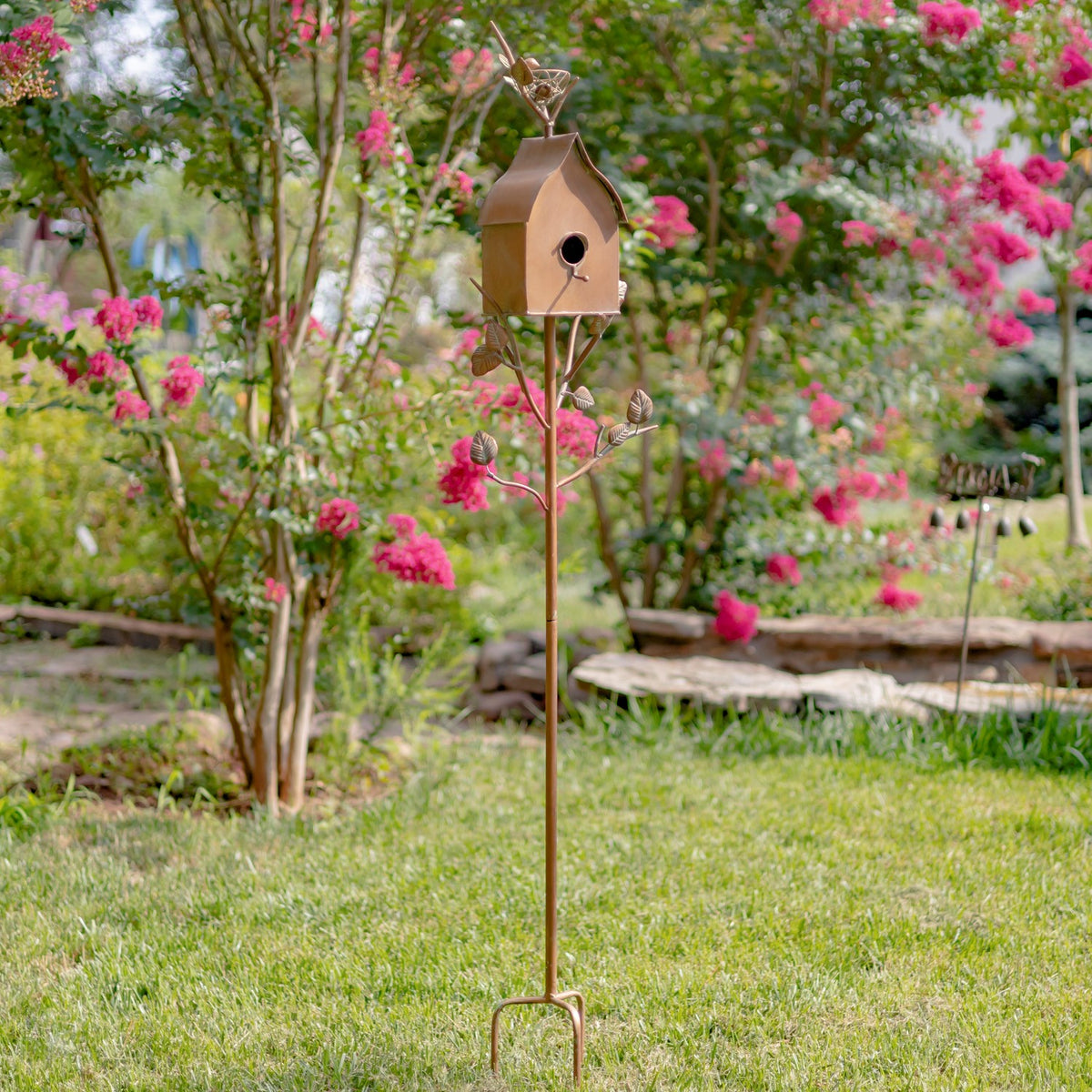 Full-length view of a birdhouse garden stake, showing its slender pole and forked ground prongs on a lawn.