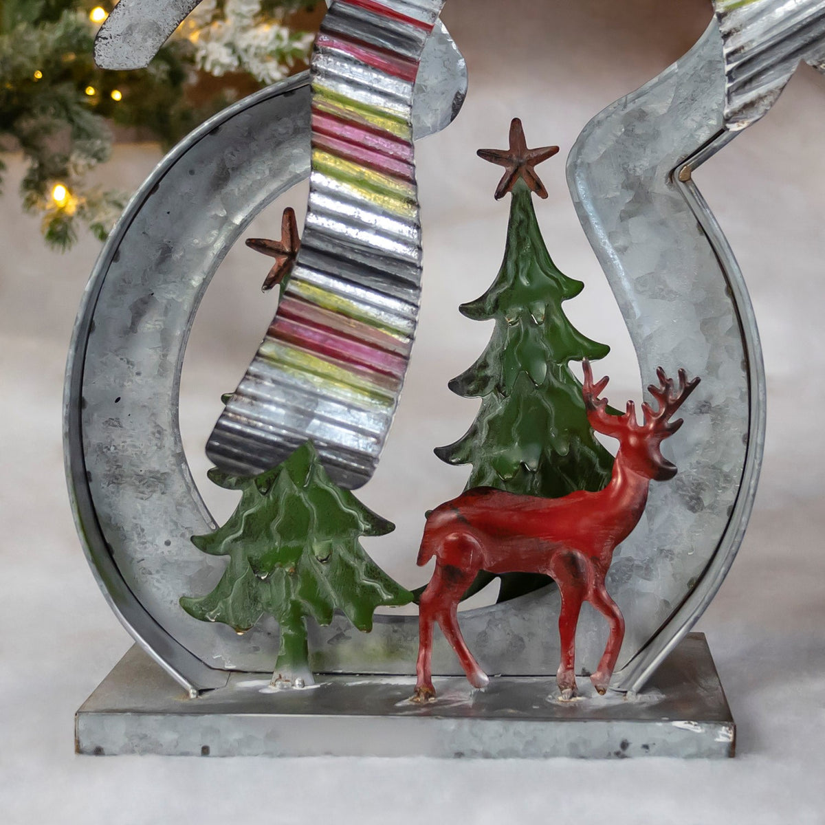 Close-up of a snowman’s curved metal body framing a green Christmas tree and red reindeer.