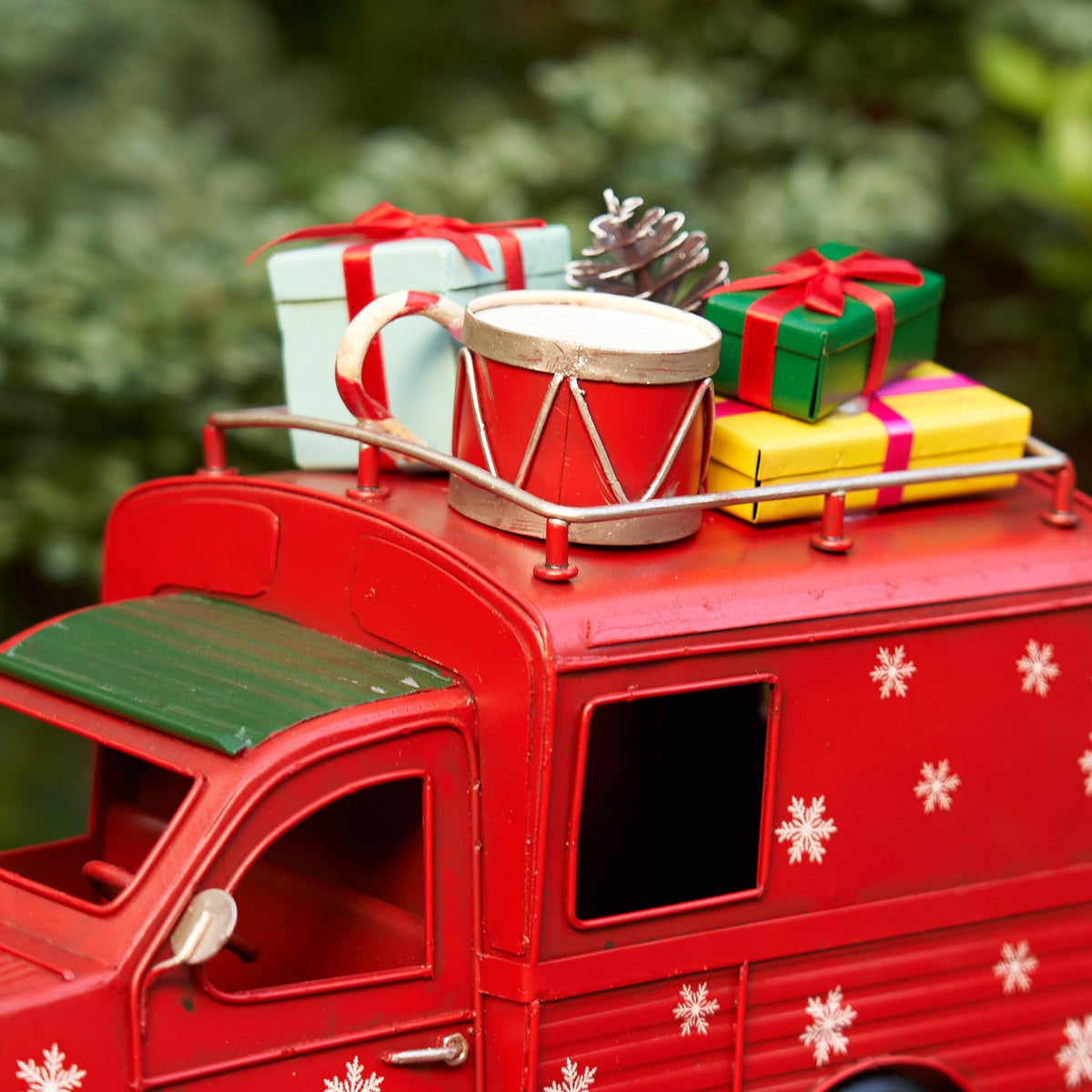 Red vintage-style truck decorated with white snowflakes, carrying wrapped gifts, a drum, and a pinecone on its roof.