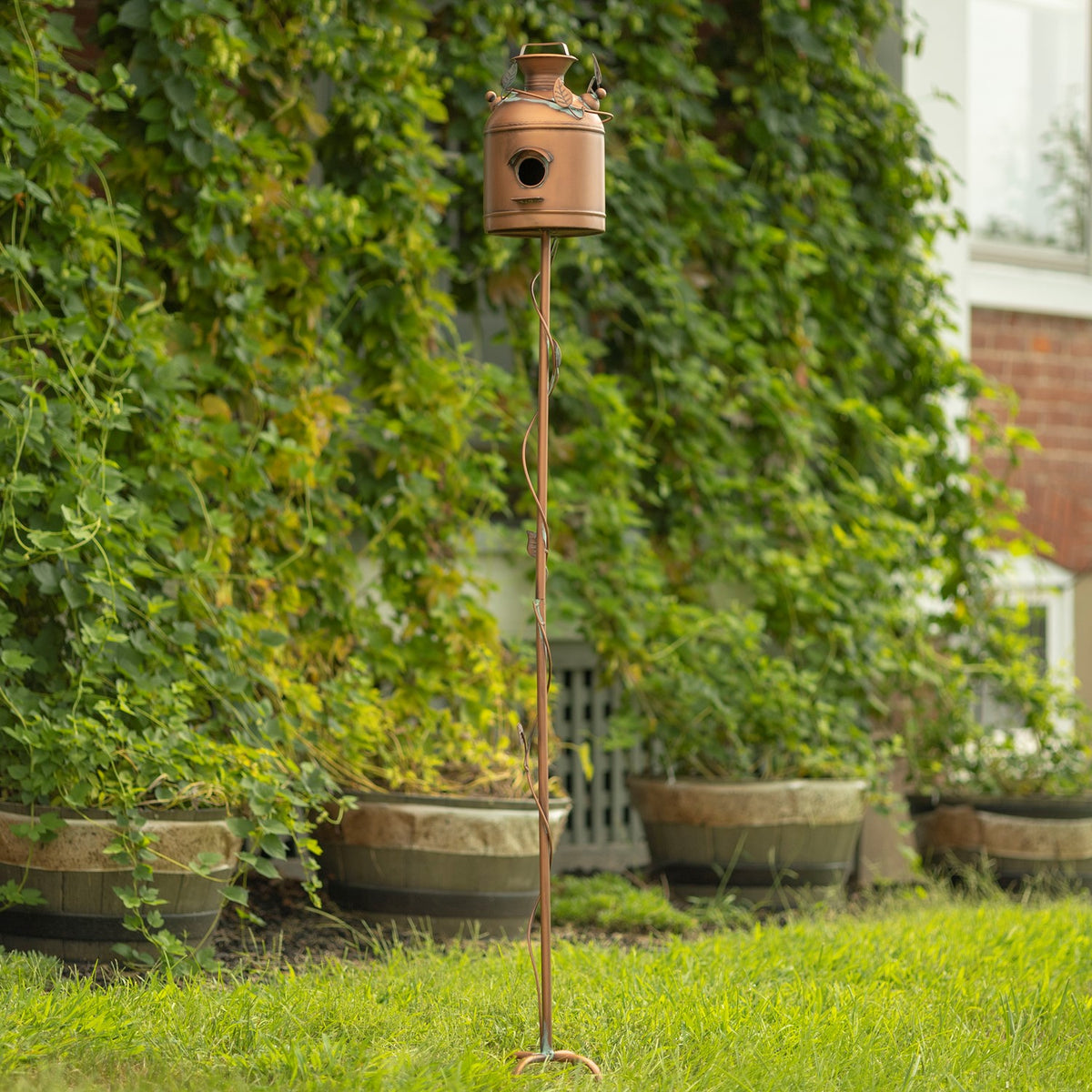 Tall birdhouse stake standing in grass beside leafy plants and garden planters.