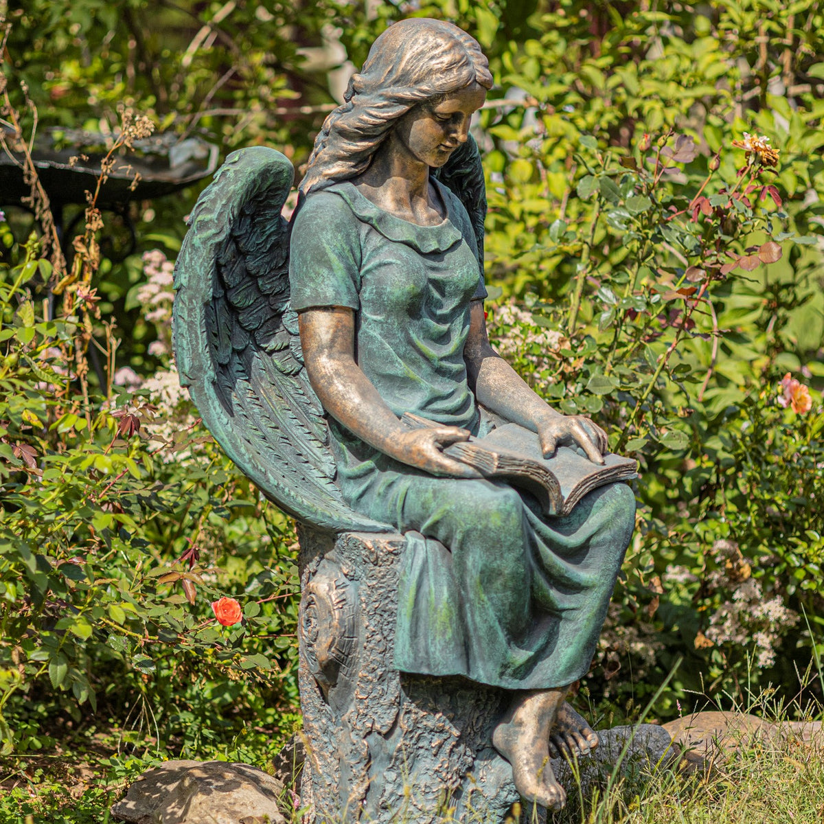 A teal-patina angel reads an open book, showing her draped dress, wings, and carved stump seat.