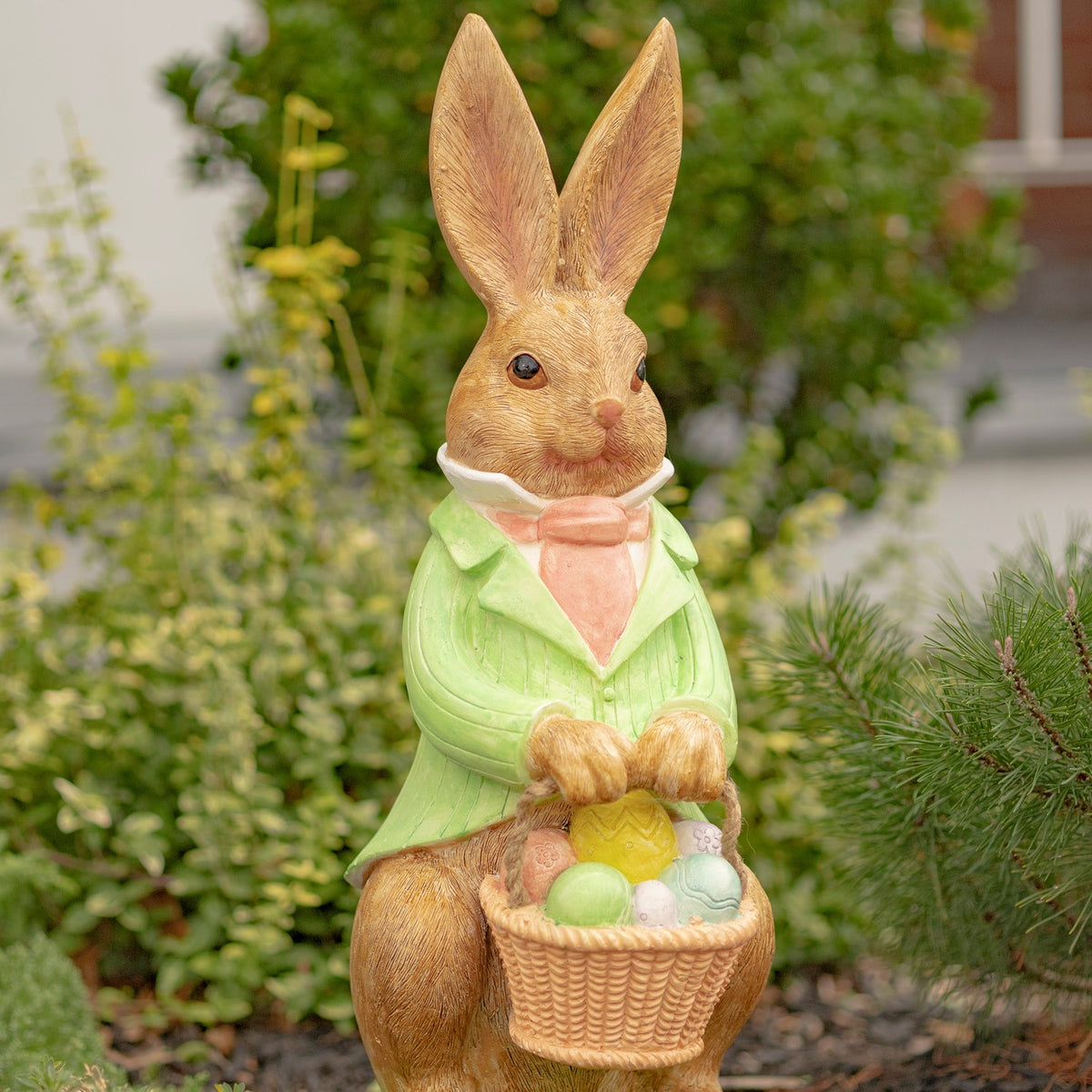 Three-quarter view of a jacketed rabbit statue holding an egg-filled basket in a garden.