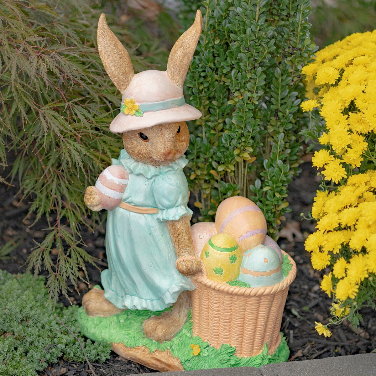 Garden rabbit statue holding a striped egg beside a woven basket filled with pastel eggs.